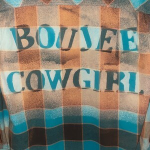May include: A blue and brown plaid flannel shirt with the words "BOUJEE COWGIRL" printed on the back in white paint.