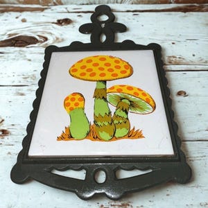 May include: A decorative trivet featuring a ceramic tile with a mushroom design. The mushrooms have orange and yellow spotted caps and green stems. The tile is set in a black, ornate metal frame with a handle.