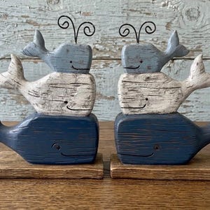 May include: Two decorative stacks of three carved wooden whales, each featuring a light blue, white, and dark blue finish. Each whale has a painted eye, a simple curved mouth, and a black wire spout on top, all mounted on a rectangular wooden base.