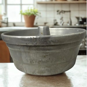 May include: A vintage, gray metal bundt pan with a central tube. The pan has a fluted design and visible text that reads "RE KAI PAN O CAK". The pan is sitting on a reflective surface, with a blurred kitchen background.