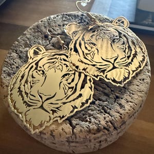 May include: Gold-colored tiger face earrings. The earrings are cut out in the shape of a tiger's face, with detailed features. The earrings are hanging on a wooden surface.