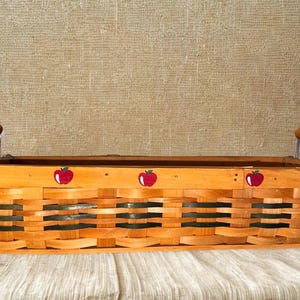 May include: A rectangular woven basket with a light brown finish and black accents. The basket features red apple designs along the top edge and wooden handles with metal supports. The basket is sitting on a light-colored surface.