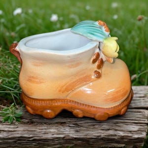 May include: A ceramic boot-shaped planter in shades of tan and brown, with a light blue and yellow floral accent. The planter sits on a weathered wooden surface against a backdrop of green grass.