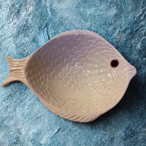 May include: A ceramic dish shaped like a fish with a glossy, light cream-colored finish. The surface features embossed, overlapping scale patterns across the body and a single, dark brown circular eye detail near the pointed snout area.