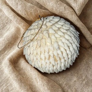 May include: A spherical, off-white decorative object covered in small, white seashells. It has a gold-colored chain and loop for hanging. The background is a textured, tan fabric.