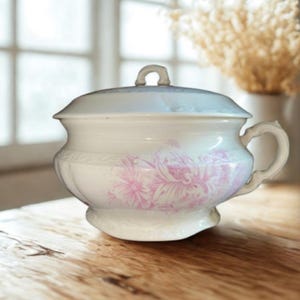 May include: A white antique chamber pot with a lid and handle. The pot features a pink floral design around the body. It is sitting on a wooden table near a window.