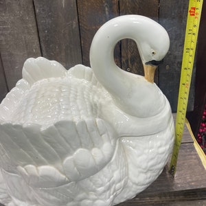 May include: A white ceramic swan-shaped serving dish with a detailed feathered design. The dish is closed, with the swan's head tucked into its body.