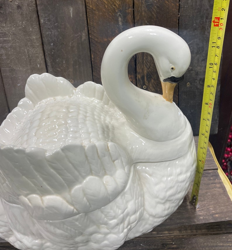 May include: A white ceramic swan-shaped serving dish with a detailed feathered design. The dish is closed, with the swan's head tucked into its body.