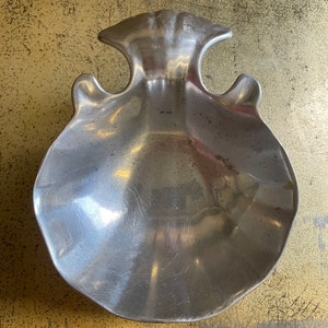 May include: A silver-colored, scallop-shaped dish with two curved handles. The dish has a textured surface and a shallow bowl. The design is reminiscent of a seashell. This decorative piece could be used as a serving dish or a decorative accent.