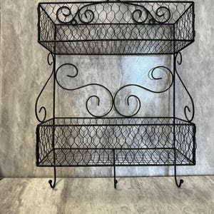 May include: A black metal wall shelf featuring two rectangular storage baskets constructed from wire mesh. The frame includes decorative scrollwork accents and three hooks attached to the bottom edge for hanging items. The structure is symmetrical.