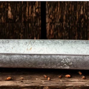 May include: A rectangular galvanized metal tray with curved handles. The tray has a distressed finish and is elevated on small feet. The background is a dark wooden surface, and the tray is likely used for decorative purposes.