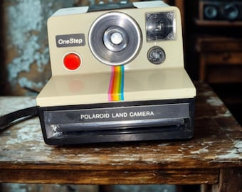 Puede incluir: Una cámara Polaroid OneStep Land vintage de color crema con una franja de arcoíris. La cámara tiene una lente negra y un botón rojo. Las palabras "OneStep" y "Polaroid Land Camera" están impresas en la cámara.