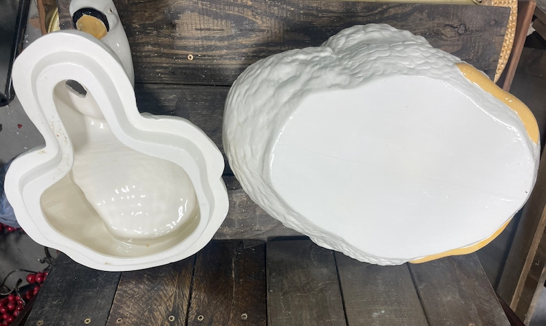 May include: A white ceramic goose cookie jar with a yellow beak and feet. The jar is open and the inside is visible. The jar is sitting on a wooden surface.