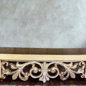 May include: A gold-painted, ornate wooden shelf with a textured top and intricate scrollwork detailing. The shelf has a rectangular shape with a beveled edge and is supported by decorative legs. The design is classical and elegant.