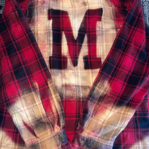 May include: Red and black plaid flannel shirt with a large, bleached-out "M" on the back. The shirt has long sleeves and a distressed, vintage look, with faded areas and a worn appearance.