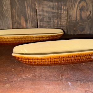 May include: Two ceramic corn-shaped serving dishes. The dishes have a textured, brown exterior resembling corn kernels and a smooth, light yellow interior. The dishes are placed on a brown surface against a wooden background.