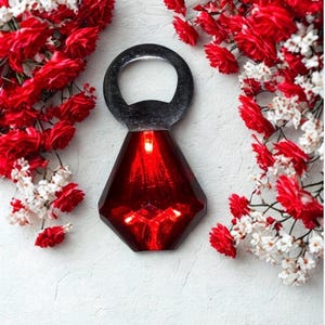 May include: A red, faceted bottle opener with a silver loop handle. The opener is surrounded by red and white flowers against a white background. The red portion of the opener has a geometric design.