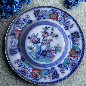 May include: A white and blue decorative plate with a floral design. The plate features a central floral arrangement with a blue vase and is surrounded by a blue border with floral and leaf patterns. The rim has a gold trim.
