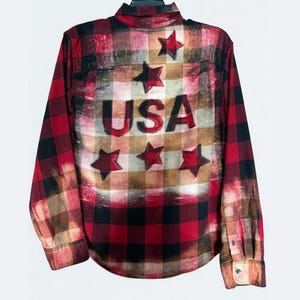 May include: Red, black, and brown plaid long-sleeve shirt with the word "USA" and star accents. The shirt has a button-down front and cuffs. The back of the shirt features a distressed design.