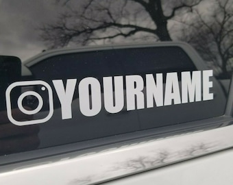 May include: White Instagram logo sticker with the text "YOURNAME" in a bold, sans-serif font. The sticker is applied to a dark window, with a blurred outdoor scene visible in the background. The sticker is likely for a car or other vehicle.