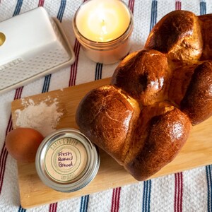 May include: A freshly baked brioche loaf on a wooden chopping board with a jar of flour, a butter dish, and a lit candle. The jar has a label that says "Fresh Baked Bread".