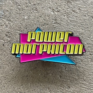 May include: A rectangular enamel pin with the text "POWER MORPHILON" in yellow, outlined in black. The pin has a pink background with blue and yellow accents. The pin is set against a gray, textured surface.