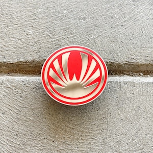 May include: A red and silver circular pin with a stylized sunburst design.