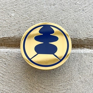 May include: A round, gold-colored pin with a navy blue design in the center. The design is a stylized, stacked shape within a gold border. The pin is set against a textured, gray surface.