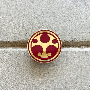May include: A gold-colored pin with a red background. The pin features a stylized symbol with three points.