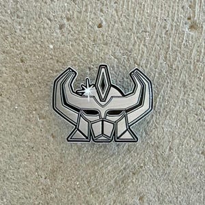 Op de afbeelding: Een zilverkleurige metalen emaille speld in de vorm van een gestileerde gehoornde helm of masker. Het ontwerp heeft scherpe, hoekige lijnen, een centrale ruitvormige kam en een klein vierpuntig steraccent op de linkerhoorn, omlijnd in zwart.