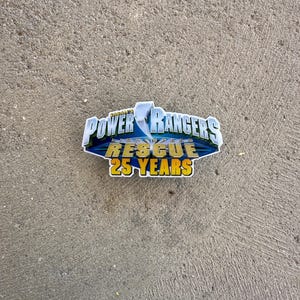 Puede incluir: Una pegatina de Power Rangers Rescue 25 Years. La pegatina presenta el logotipo de Power Rangers en azul, blanco y dorado, con las palabras "Power Rangers Rescue" y "25 YEARS" en un lugar destacado. La pegatina está sobre un fondo gris.