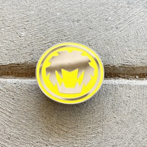 May include: A yellow and silver enamel pin with a lion's head design.