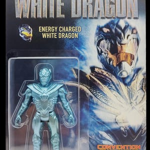 May include: A blue and silver action figure of a Power Ranger in a white dragon suit. The figure is packaged in a plastic blister pack with a blue and silver background. The packaging features the text "Legend of the White Dragon" and "Energy Charged White Dragon". The packaging also includes the text "Convention Exclusive" and "Power Morphincon".