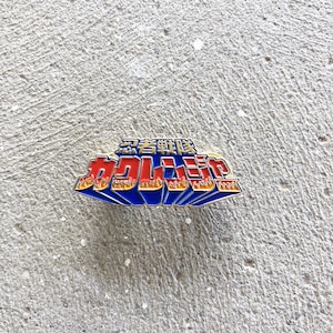 May include: A rectangular enamel pin with Japanese text in red, yellow, and blue. The text is in a stylized, 3D font with a flame-like design. The pin is set against a textured, gray concrete surface.