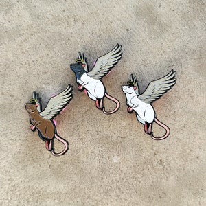 May include: Three enamel pins featuring cartoon rats with angel wings and halos. The rats are black, brown, and white.