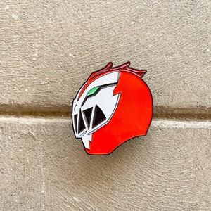 May include: Red and white enamel pin of a helmet with a green visor and black details. The helmet has a jagged design on the side.