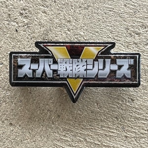 May include: A black and silver pin with a yellow V shape and the text "Super Sentai Series" in Japanese.