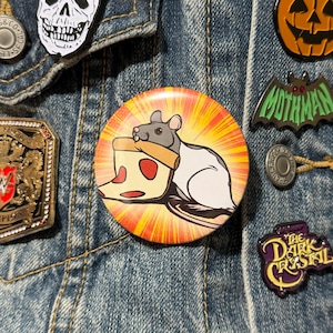 Puede incluir: Un botón redondo con un diseño de rata de dibujos animados sosteniendo una porción de pizza. La rata es gris y blanca, la porción de pizza es roja, amarilla y marrón. El botón tiene un fondo de ráfaga amarillo y naranja.