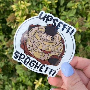 May include: A white sticker with a black outline and the text "Upset Spaghetti" in black. A cartoon snake wearing a black hat is in the center of the sticker, curled up on a plate of spaghetti.