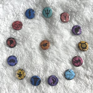 Könnte beinhalten: Ein Satz von 12 runden Knöpfen mit verschiedenen Symbolen und Farben. Die Knöpfe sind in einem Kreis auf einer weißen, flauschigen Oberfläche angeordnet. Die Symbole umfassen einen Blitz, einen Dreizack, einen Kreis mit einem Symbol darin, eine Weinrebe, eine Silhouette eines Frauenkopfes, ein Pferd und einen Wagen, eine Leier, einen Vogel mit ausgebreiteten Flügeln, eine Lotusblume, einen Amboss, einen Hermesstab und einen zunehmenden Mond mit einem Vogel.