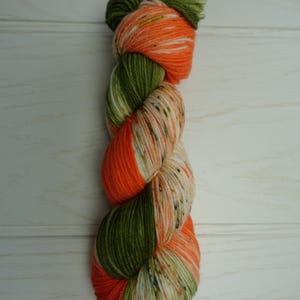 Könnte beinhalten: Ein Knäuel farbenfroher Wolle in Orange-, Grün- und Cremetönen. Das Garn ist gezwirnt und scheint eine Mischung aus verschiedenen Fasern zu sein. Die Farben sind lebendig und das Garn hat ein gesprenkeltes Aussehen, geeignet für Strick- oder Häkelprojekte.