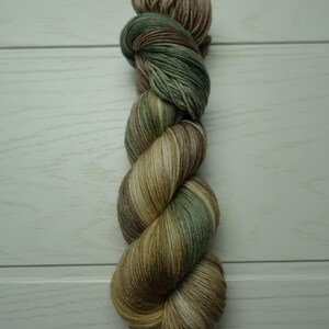 Könnte beinhalten: Ein Strang handgefärbter Wolle in Erdtönen. Das Garn ist eine Mischung aus Braun, Grün und Beige, wodurch ein mehrfarbiger Effekt entsteht. Das Garn ist zu einem Strang gezwirnt, bereit für Strick- oder Häkelprojekte.