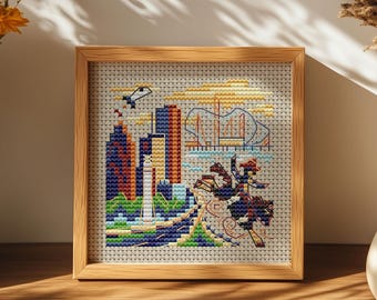May include: A framed cross-stitch artwork depicting a cityscape with skyscrapers, a bridge, and figures. The artwork features a wooden frame and is displayed on a wooden surface. The color palette includes blues, oranges, and greens.