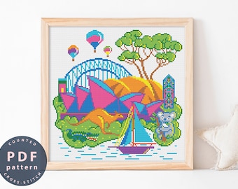 May include: A counted cross stitch pattern featuring a colorful illustration of the Sydney Opera House, a sailboat, a kangaroo, a crocodile, and hot air balloons. The pattern is framed in a light wood frame.