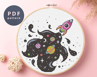 May include: Cross stitch pattern featuring a black and white space scene with a pink and green rocket shooting out of the center. The space scene includes planets, stars, and a crescent moon.