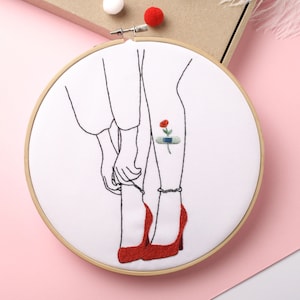 May include: Embroidered hoop art featuring a line drawing of a person's legs with red shoes and a small red flower with a bandage on the ankle.