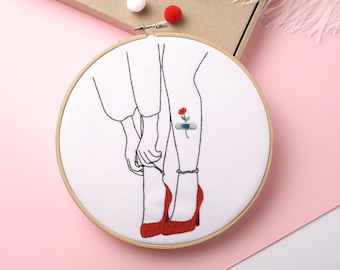 May include: Embroidered hoop art featuring a line drawing of a person's legs with red shoes and a small red flower with a bandage on the ankle.