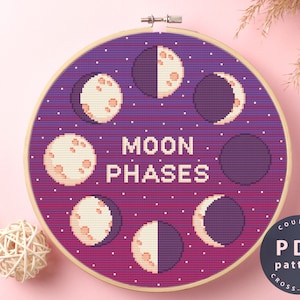 May include: A counted cross stitch pattern featuring a purple and pink gradient background with white moons in different phases. The text "MOON PHASES" is in the center of the design. The pattern is in a round embroidery hoop.