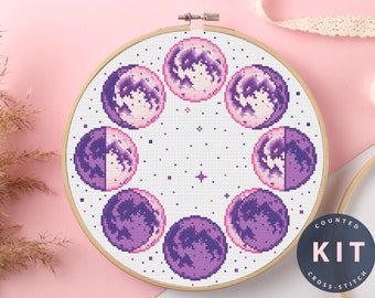 May include: A purple and white cross stitch pattern of the moon phases in a circle. The pattern is in a wooden embroidery hoop. The text "COUNTED KIT CROSS STITCH" is on a black circle.