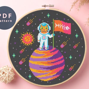 May include: Cross stitch pattern featuring a cat astronaut standing on a planet with a flag that says "Mike". The background is a dark blue space with stars and shooting stars.